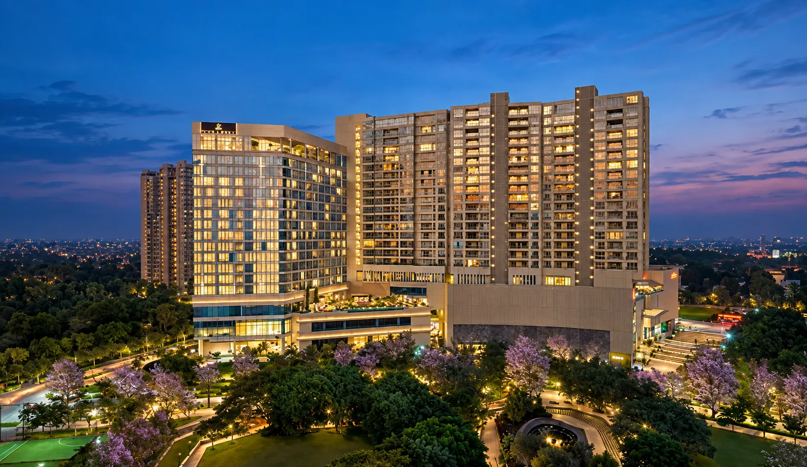 The Leela Bhartiya City hotel lit up at dusk above jacaranda trees.